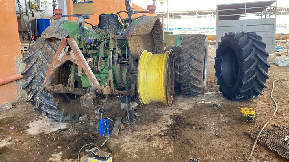 טרקטור ירוק בלי גלגל אחורי, מורם על ג'ק ותומכת, לצידו חישוק צהוב וצמיג חדש